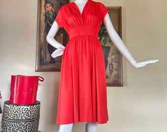 May include: A bright red dress with a V-neck and short sleeves. The dress has a fitted waist and a flowing skirt. The dress is displayed on a mannequin. A red hat box and two leopard print hat boxes are in the background.