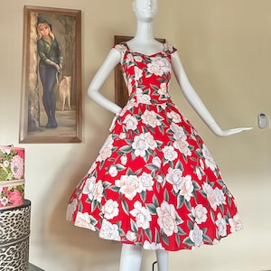 May include: A sleeveless red dress featuring a repeating floral pattern of white and pale pink roses with dark green leaves. The dress has a fitted bodice with a sweetheart neckline and a full, flared skirt that reaches below the knee.