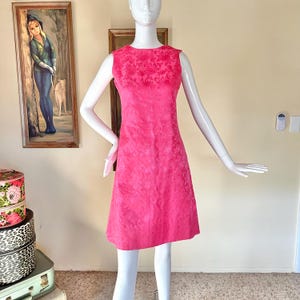 May include: A sleeveless, knee-length, hot pink dress on a mannequin. The dress features a textured fabric and a floral pattern on the bodice. The dress has a simple, classic silhouette.
