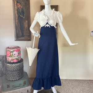 May include: A floor-length navy blue dress with a ruffled hem, paired with a short-sleeved white bolero jacket featuring ruffled edges and a central white floral applique. The ensemble is displayed on a mannequin holding a small white parasol.