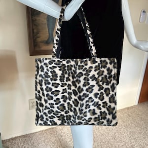 May include: A rectangular tote bag with a leopard print pattern in shades of black, brown, and cream. The bag has a structured shape with a black base and matching handles. The bag is held by a mannequin.