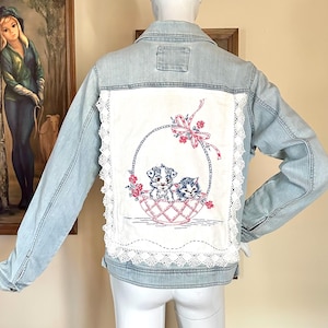 May include: Light blue denim jacket with a white embroidered panel on the back. The panel features a basket with a puppy and kitten, accented with pink flowers and a bow. The panel is trimmed with white lace.
