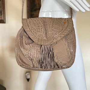 May include: A tan leather shoulder bag with a flap closure and a long strap. The bag features a textured pattern, a tassel, and a small gold-colored square emblem. The bag is displayed on a white mannequin.