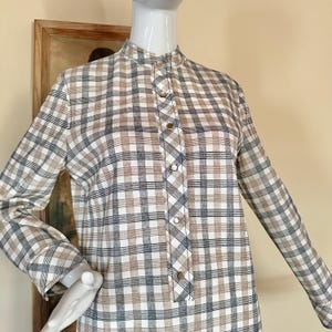 May include: A long-sleeved shirt featuring a plaid pattern in shades of white, tan, and dark gray. The shirt has a high collar and a central vertical placket with five visible metallic buttons running down the front center of the garment.