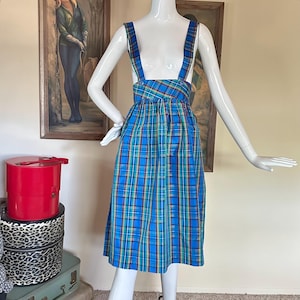 May include: A blue plaid suspender dress on a mannequin. The dress features a vibrant pattern of blue, green, and yellow lines. The suspenders are attached to the bodice and cross in the back. The dress is knee-length and has a gathered waist.