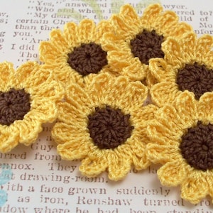 May include: Six crocheted yellow sunflower embellishments with brown centers. The sunflowers are arranged in a cluster on a white background.