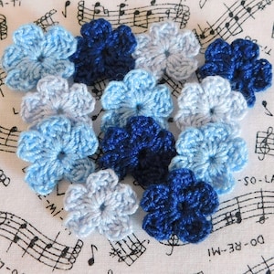 May include: A collection of handmade crochet flowers in various shades of blue, including light, medium, and dark tones. The flowers are arranged on a white fabric with musical notes printed on it. The flowers are a charming craft item.