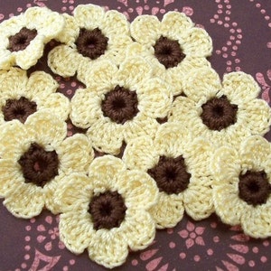 May include: A collection of crocheted flowers with a daisy-like design. The petals are a creamy yellow, and the centers are a deep brown. The flowers are handmade and appear to be made of yarn.