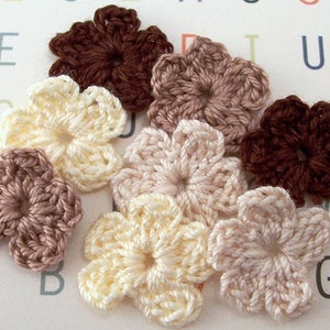 May include: A collection of seven crocheted flower embellishments in various shades of brown, beige, and cream. The flowers are all the same size and shape, with five petals each.