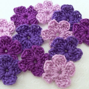 May include: A collection of crocheted flower embellishments in shades of purple and lavender. The flowers are made with a soft yarn and have a delicate, feminine look.