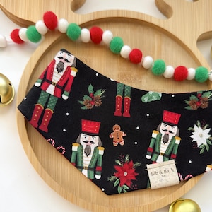 May include: A black pet bandana with a festive Christmas theme. The bandana features a repeating pattern of nutcrackers in red and green uniforms, gingerbread men, poinsettias, and snowflakes. The bandana is on a wooden tray with a red, white, and green pom-pom garland.