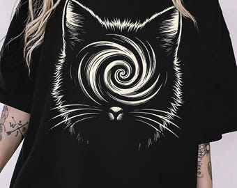 May include: Black t-shirt featuring a stylized cat face with a spiral design in the center. The cat's face is outlined in white, creating a striking contrast against the black fabric. The shirt has short sleeves and a relaxed fit.