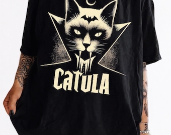 Puede incluir: Camiseta negra con un diseño estilizado de gato con colmillos de vampiro y un símbolo de murciélago. La palabra "CATULA" está impresa debajo. La camiseta tiene un corte holgado y es de tejido suave.