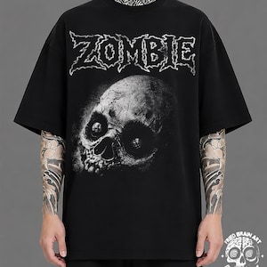 May include: A black short-sleeve t-shirt featuring the word ZOMBIE in a jagged white font across the chest. Below the text is a detailed, realistic graphic of a human skull with deep, dark eye sockets. A small logo with a skull and the text FRIED BRAIN ART is visible in the bottom right corner.
