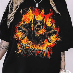 May include: Black t-shirt featuring a graphic of three snarling black dogs with red eyes, surrounded by flames. The word "Feral" is displayed in a gothic font. The design is a bold, eye-catching statement.