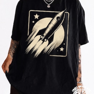 May include: Black t-shirt featuring a retro rocket ship graphic. The design includes a rocket blasting off against a cream-colored moon and stars, set within a square frame. The text "FRIED BRAIN ART" is visible at the bottom.