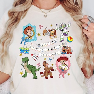 May include: Cream-coloured t-shirt featuring a colourful graphic of characters from the animated film "Toy Story." The design includes Woody, Buzz Lightyear, and other characters, along with the text "You've Got a Friend in Me."