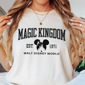 May include: Ivory-colored t-shirt with the text "MAGIC KINGDOM EST. 1971 WALT DISNEY WORLD" in black. The design includes a silhouette of Mickey Mouse ears and a castle. The shirt is a casual wear item.