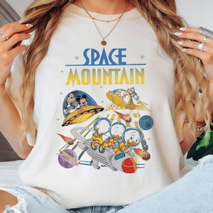 以下が含まれることがあります： アイボリー色のTシャツ。ディズニーキャラクターが宇宙をテーマにしたカラフルなグラフィック。デザインには「SPACE MOUNTAIN」の文字が青と黄色で描かれ、ミッキーマウス、ドナルドダック、その他のキャラクターが宇宙を舞台に描かれています。
