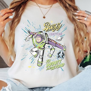 May include: Ivory-colored t-shirt featuring a graphic of Buzz Lightyear from Toy Story. The design includes the text "Buzz Lightyear" and "To Infinity and Beyond" in a colorful, cartoon style. The shirt is a crew neck and has short sleeves.