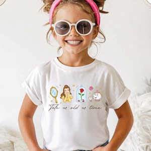 Può includere: T-shirt bianca con un'illustrazione colorata dei personaggi e degli oggetti de La Bella e la Bestia. Il design include uno specchio, Belle, una rosa in una teca di vetro e Mrs. Potts e Chip. Il testo "Tale as old as time" è scritto sotto.