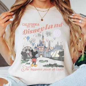 May include: Ivory-colored t-shirt featuring a Disneyland graphic. The design includes the words "California Est. 1955 Disneyland" in pink script, along with an illustration of the castle, Mickey Mouse, and other park elements. The phrase "The happiest place on Earth" is also included.