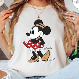 Puede incluir: Camiseta de color marfil con un dibujo de Minnie Mouse. El personaje de dibujos animados lleva una falda roja de lunares y zapatos amarillos. El logotipo de Disney est&aacute; impreso debajo. La camiseta tiene cuello redondo y mangas cortas.