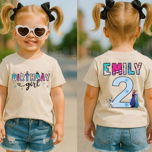 May include: A tan t-shirt with the text "Birthday girl" and a graphic of characters. The back of the shirt has the name "Emily" and the number "2" with cartoon characters. The child is wearing denim shorts.