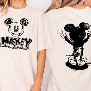 May include: Cream-colored t-shirt featuring a black and white Mickey Mouse graphic. The front of the shirt displays Mickey's face with the word "Mickey" below. The back shows Mickey with his arms raised in a rock-and-roll gesture.