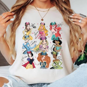 Op de afbeelding: Ivoor kleurig T-shirt met illustraties van Disney-prinsessen en hun handtekeningen. De prinsessen zijn afgebeeld in hun iconische outfits, met details zoals strikken en kronen. Het woord "IVORY" staat onderaan.