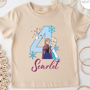 May include: A beige short-sleeved t-shirt with a blue number 4, snowflakes, and cartoon characters Anna and Elsa. The name "Scarlet" is written in a cursive font below the characters. The shirt is designed for a child.