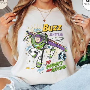 K&ouml;nnte beinhalten: Elfenbeinfarbenes T-Shirt mit einem Buzz Lightyear-Motiv und dem Text "Buzz Lightyear" und "To Infinity and Beyond". Das Design enth&auml;lt gr&uuml;ne, lila und wei&szlig;e Elemente. Das T-Shirt hat einen Rundhalsausschnitt.