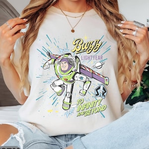 Może przedstawiać: Kremowa koszulka z grafiką Buzza Astrala w dynamicznej pozie. Projekt zawiera napisy "Buzz Lightyear" i "To Infinity and Beyond" w kolorowej czcionce w stylu komiksowym. Jest to element odzieży codziennej.