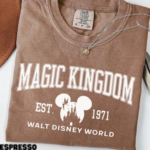 May include: Espresso-colored t-shirt with "MAGIC KINGDOM" in white block letters. Below the text is a white graphic of Mickey Mouse ears and a castle, with "EST. 1971" and "WALT DISNEY WORLD" below. A silver necklace with a pendant rests on the shirt.