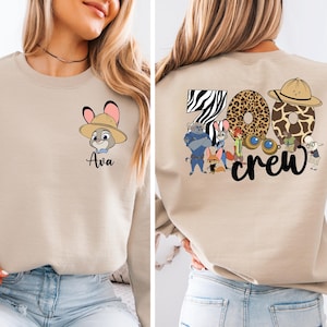 Puede incluir: Sudadera beige con un conejo de dibujos animados con sombrero y el nombre "Ava". La parte trasera tiene "Zoo Crew" con estampados de animales y personajes de dibujos animados. Una parte superior informal y divertida.