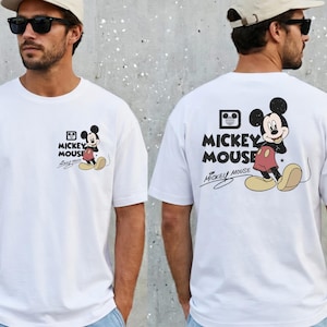 May include: White t-shirt featuring Mickey Mouse. The front of the shirt has a small graphic of Mickey Mouse with the words "Mickey Mouse" and a signature. The back of the shirt has a larger Mickey Mouse graphic and the words "Mickey Mouse".