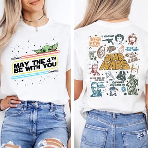 以下が含まれることがあります： 白いTシャツ。前面には、ライトセーバーと「MAY THE 4TH BE WITH YOU」の文字と、ベビーヨーダのグラフィック。背面にはスターウォーズのキャラクターと引用句が描かれています。カジュアルウェアアイテムです。