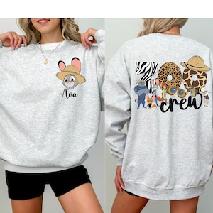Puede incluir: Sudadera gris claro con cuello y pu&ntilde;os blancos. La parte delantera presenta un personaje de dibujos animados con un sombrero y el nombre "Ava". La parte posterior muestra "Zoo Crew" con letras y personajes tem&aacute;ticos de animales.