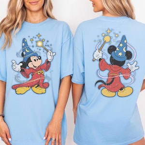 Könnte beinhalten: Hellblaue T-Shirts mit einem Retro-Design von Mickey Mouse als Zauberer, der einen Zauberstab mit Sternen hält. Vorder- und Rückseite des Shirts sind abgebildet. Mickey trägt eine rote Robe und einen blauen Hut mit Sternen.