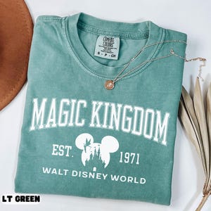 May include: A light green t-shirt with the words "MAGIC KINGDOM" in white block letters. Below the text is "EST. 1971" and "WALT DISNEY WORLD". A gold necklace with a pendant rests on the shirt. The shirt is labeled "LT GREEN".