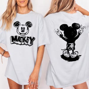 May include: Light blue t-shirt featuring a black and white Mickey Mouse graphic on the front and back. The front design includes Mickey's face and the word "Mickey". The back design shows Mickey with his arms raised in a rock and roll gesture.
