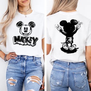 May include: White t-shirt featuring a black and white graphic of Mickey Mouse on the front with the word "Mickey" in a block letter style. The back of the shirt shows a black and white illustration of Mickey Mouse with his hands up in a rock and roll gesture.