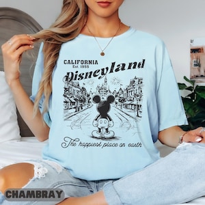 May include: Light blue t-shirt with a black and white Disneyland graphic. The graphic features Mickey Mouse in front of a street scene with the text "California Est. 1955 Disneyland The happiest place on earth."