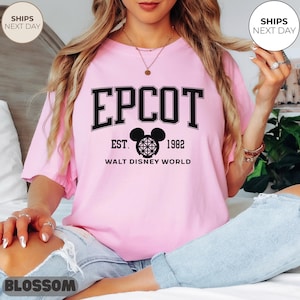 May include: A light pink t-shirt with the word "EPCOT" in black block letters. Below the word is a black Mickey Mouse head design and the text "EST. 1982 WALT DISNEY WORLD". The shirt has short sleeves and a crew neck.
