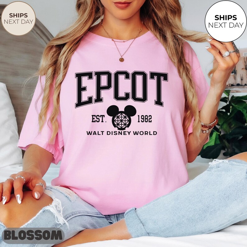 May include: A light pink t-shirt with the word "EPCOT" in black block letters. Below the word is a black Mickey Mouse head design and the text "EST. 1982 WALT DISNEY WORLD". The shirt has short sleeves and a crew neck.