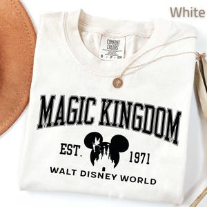 May include: Cream-colored t-shirt with the words "MAGIC KINGDOM" in black block letters. Below the text is a silhouette of Mickey Mouse ears and a castle, with the text "EST. 1971 WALT DISNEY WORLD". A gold necklace is draped across the shirt.