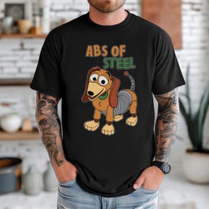 K&ouml;nnte beinhalten: Schwarzes T-Shirt mit einem Cartoon-Dackel-Design und dem Text "ABS OF STEEL". Der Hund hat braune Ohren, eine gelbe Nase und einen gr&uuml;nen Kragen. Der K&ouml;rper des Hundes ist eine silberne Feder. Ein lustiges, verspieltes Design.