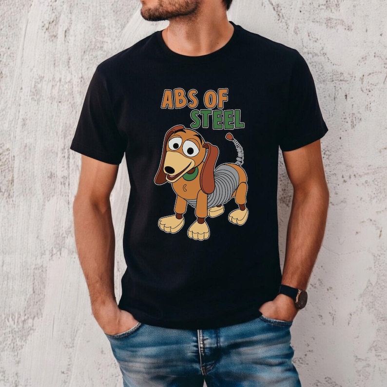 K&ouml;nnte beinhalten: Schwarzes T-Shirt mit einer Cartoon-Illustration von Slinky Dog und dem Text "ABS OF STEEL". Das Design zeigt den Federk&ouml;rper der Figur und ihren freundlichen Gesichtsausdruck. Ein lustiges, verspieltes Design.