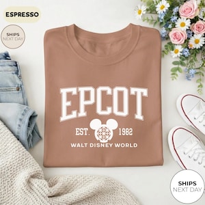 May include: Espresso-colored t-shirt with the word "EPCOT" in white block letters. Below the word "EPCOT" is the text "EST. 1982" and a Mickey Mouse head graphic. The words "WALT DISNEY WORLD" are printed below the graphic. The shirt is folded.