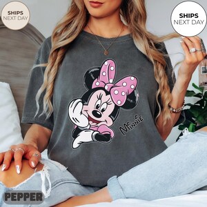 May include: Dark gray t-shirt featuring a Minnie Mouse graphic. Minnie is depicted with a pink bow with white polka dots, a pink dress, and white gloves. The word "Minnie" is written in cursive.